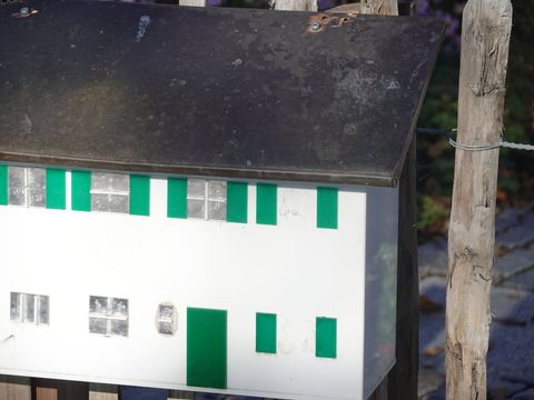 Λευκό γραμματοκιβώτιο σε σχήμα σπιτιού με πράσινα παντζούρια πάνω σε ξύλινο φράχτη