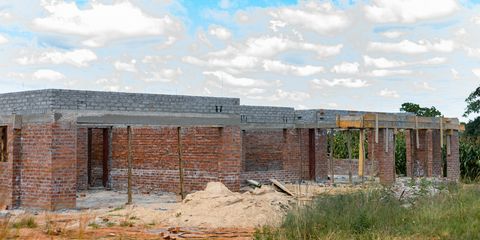 Gemauerter Rohbau eines flachen Hauses aus roten Ziegeln und Betonbalken, noch ohne Dach und Fenster, davor ein Sandhaufen, dahinter ein Maisfeld unter bewölktem Himmel.
