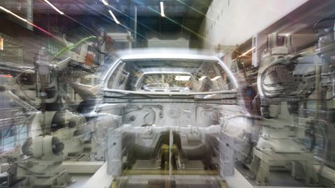 Unscharfe Langzeitbelichtung einer Autokarosserie im Rohbau, links und rechts schwenken weiße Industrieroboterarme, Lichtstreifen der Hallenbeleuchtung ziehen durchs Bild.